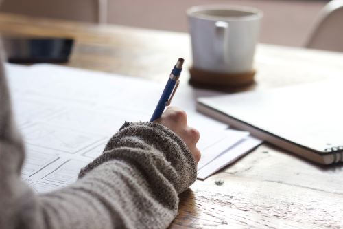 neue gewohnheiten etablieren - schreibende Person in grauem Pullover am Tisch mit einem Kugelschreiber in der Hand, über einigen Zetteln, daneben ein Notizbuch und eine Tasse